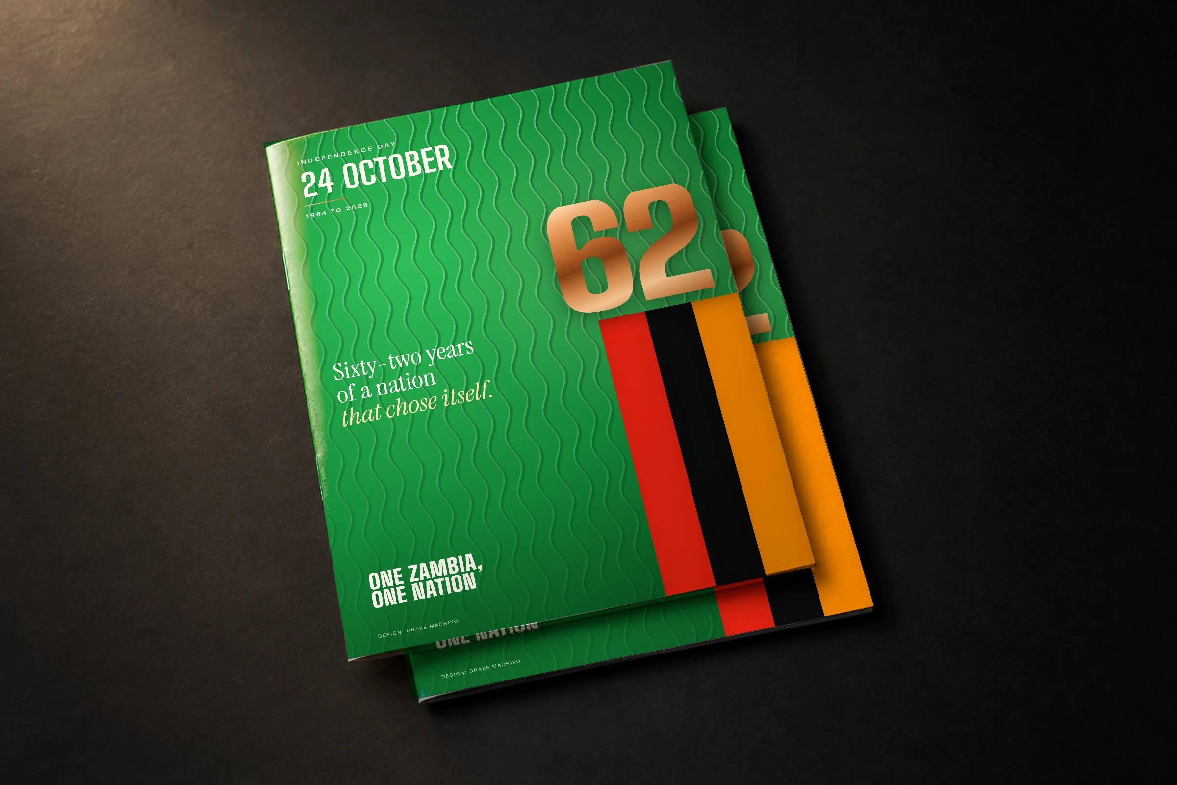 Zambia at 62 printed as two booklets on a dark desk: the green field with the falls drawn as waves, the flag stripes and a copper 62