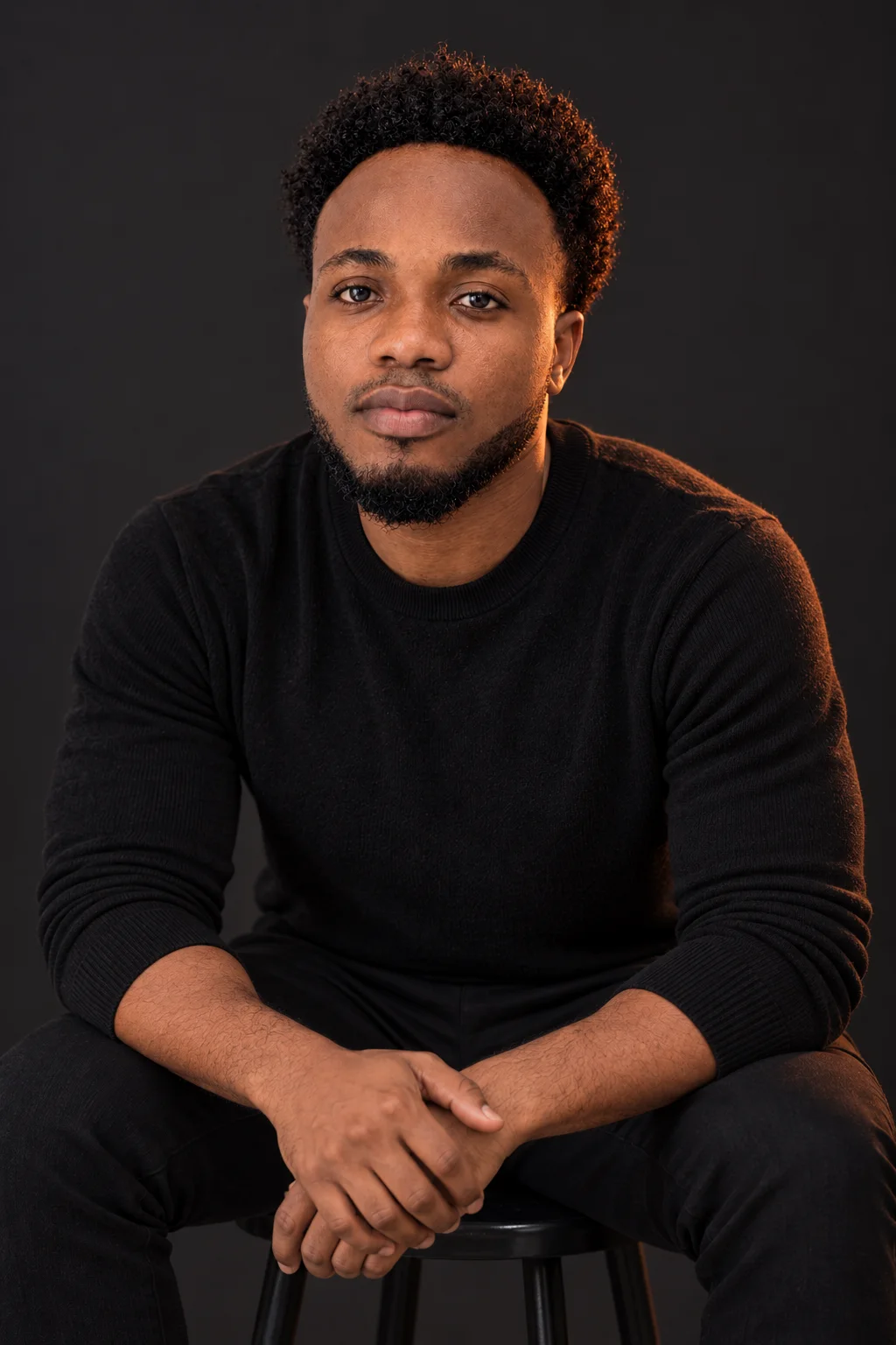 Seated on a studio stool in a black knit sweater, looking at the camera